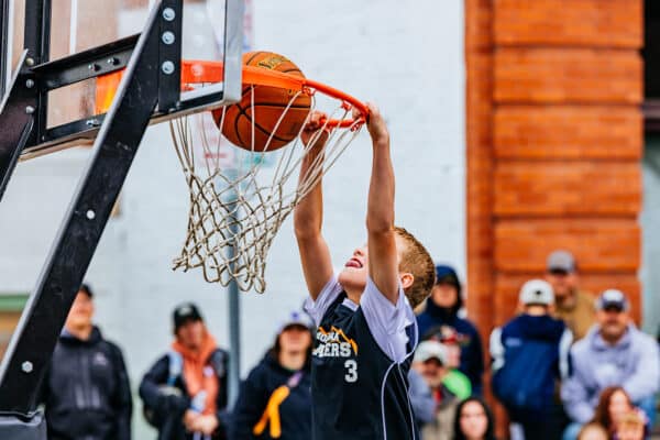 Youth basketball player finishing a layup at an outdoor street basketball event in Butte, Montana, with spectators watching.